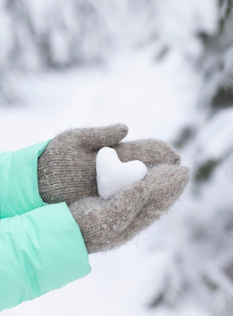 ニットの暖かいミトンを着た女性の手は、雪に覆われた森の中で雪で作られた心を持っています