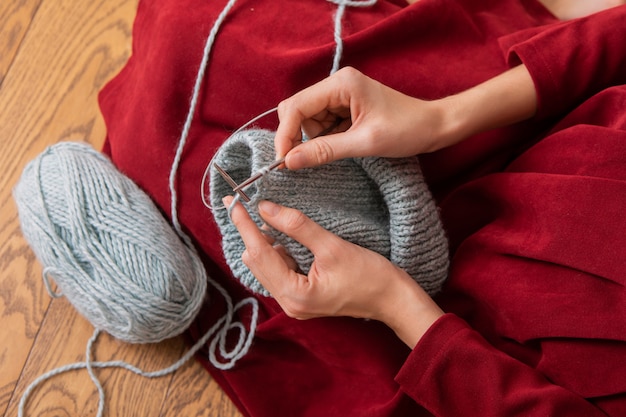 Women's hands knitting gray hat. Sitting at the wooden floor.