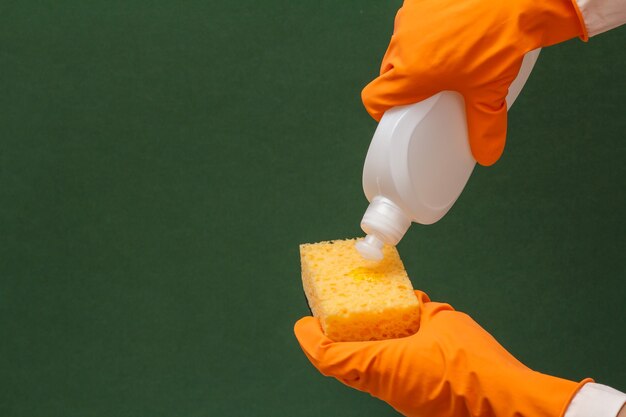 Women's hands in orange protective glove with bottle of dishwashing liquid and sponge on the green background. Washing and cleaning concept.