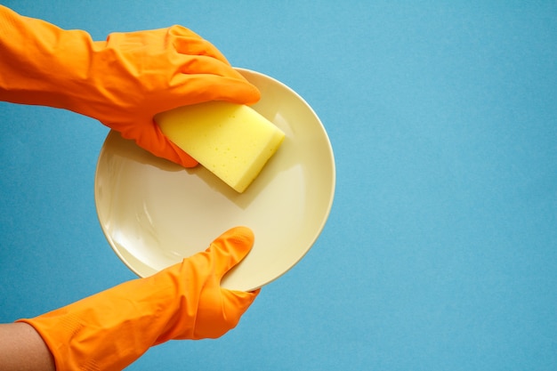 Women's hands in orange rubber gloves with plate and sponge on blue background. Washing and cleaning concept.