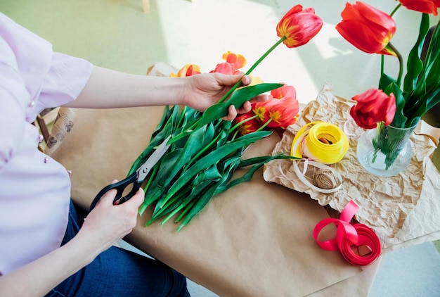 Women's hands pack a festive bouquet in wrapping paper The florist makes an assembly with red tulips in the workshop A woman at work small business or hobby