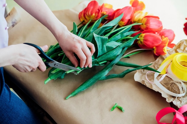 Women's hands pack a festive bouquet in wrapping paper The florist makes an assembly with red tulips in the workshop A woman at work small business or hobby