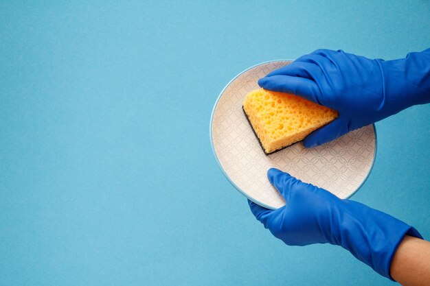 Women's hands in protective gloves with plate and sponge on blue background. Washing and cleaning concept.