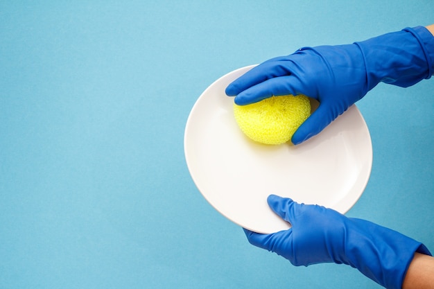 파란색 배경에 흰색 접시와 노란색 스폰지가 있는 보호 장갑을 끼고 있는 여성의 손. 세척 및 청소 개념입니다.