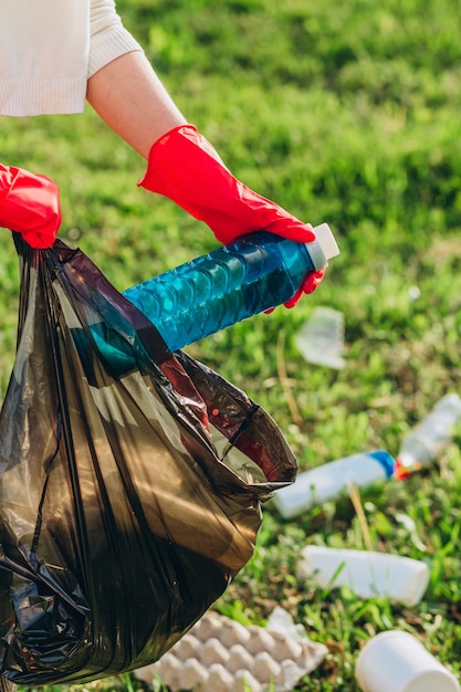 Women's hands in red rubber gloves. Woman collects trash in the bag. Volunteer scavenge garbage in the summer park. Nice progressive woman making an effort to help the environment