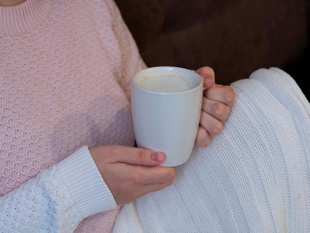 カプチーノの暖かさの快適さの白いマグカップと暖かいニットセーターの女性の手