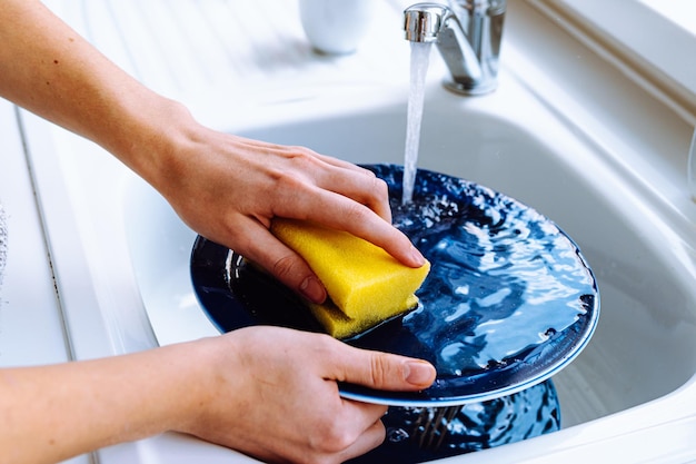 Women\'s hands wash dirty plate with sponge for dishes under\
stream of water from tap