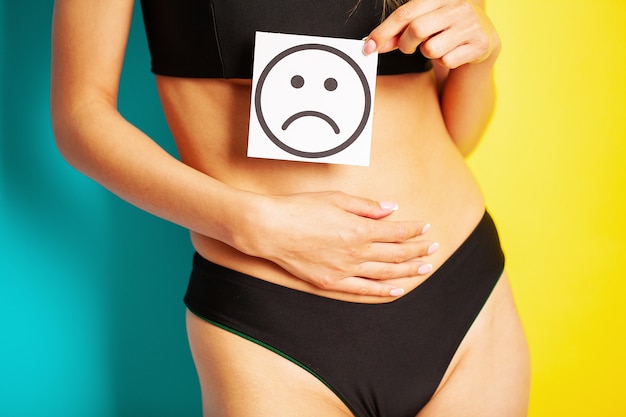 Women's health, women's body holds a card of a sad smile near the belly.