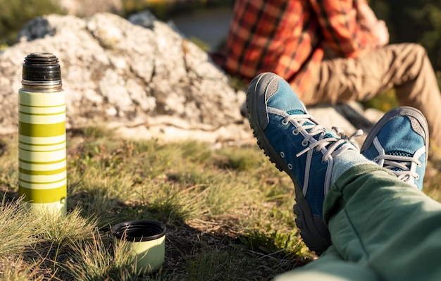 写真 ハイキングブーツを履いた女性の足は秋に山の川でリラックスし男性はサーモスからお茶を飲んでいます