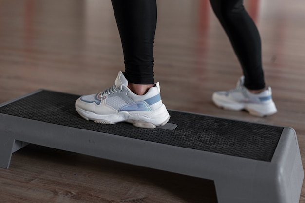 세련된 스포티 한 흰색 스니커즈의 여성 다리가 체육관의 플랫폼 계단에 서 있습니다.