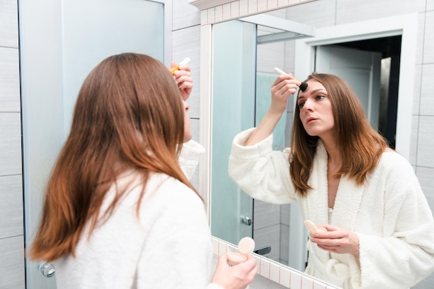 Women's morning routine A young woman in a white bathrobe puts makeup on her face with a brush