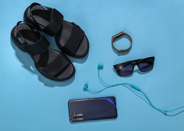 Women's sandals (smartphone, smart bracelet, headphones), sunglasses on a blue wooden background. Top view