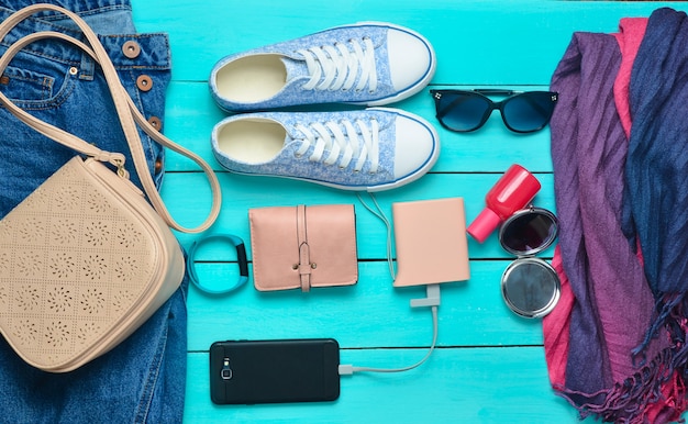 Accessori alla moda femminile, scarpe, vestiti e gadget moderni su un fondo di legno blu. jeans, borsa, scarpe da ginnastica, smartphone, braccialetto intelligente, power bank, cosmetici, occhiali da sole, sciarpa. vista dall'alto.