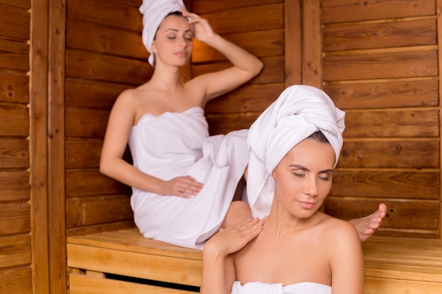 Women in sauna. Two attractive women wrapped in towel relaxing in sauna and keeping eyes closed