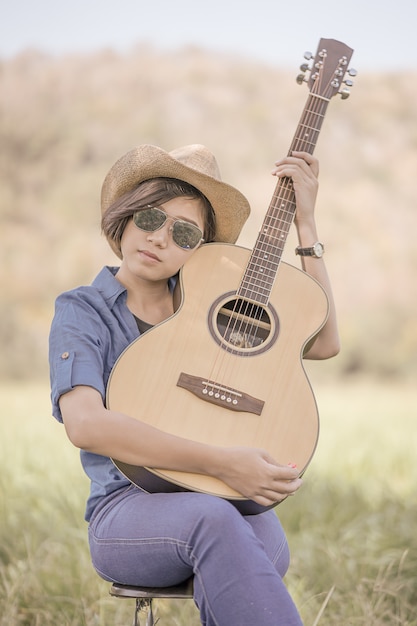 Women short hair wear hat and sunglasses sit playing guitar in grass field 