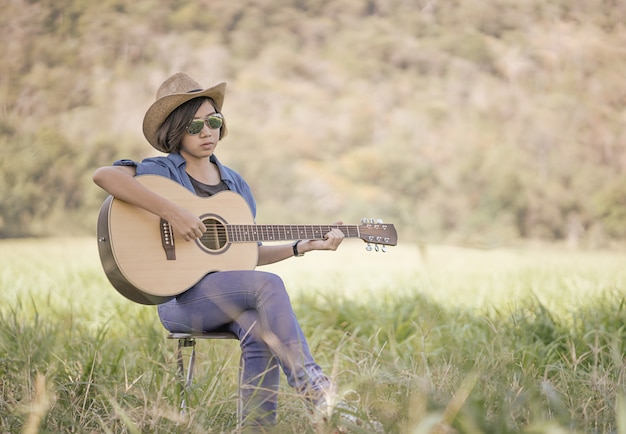 Women short hair wear hat and sunglasses sit playing guitar in grass field 