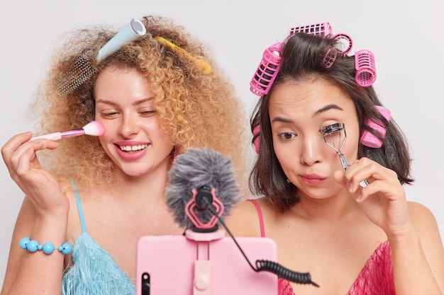 Women show how to do professional makeup stand in front of\
smartphone webcam apply face powder with brush curl eyelashes with\
curlers make hairstyle record video blog talk to followers