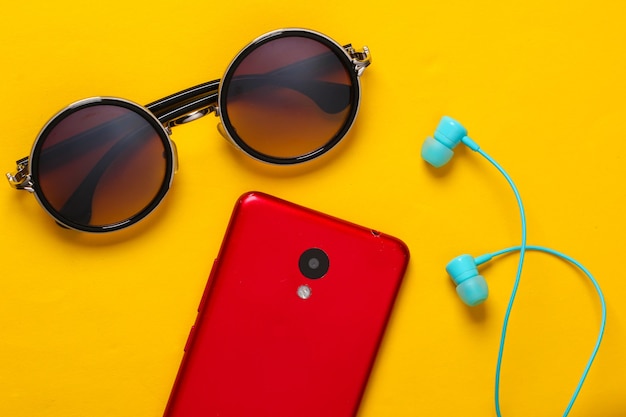 Womens accessories and gadgets on a yellow. Earphones, stylish round sunglasses, smartphone on a yellow