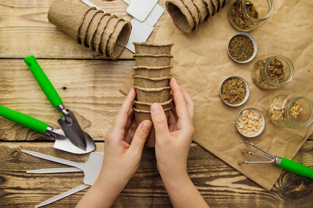 Womens handen houden turf potten bovenaanzicht van zaden en tuingereedschap op een houten achtergrond groeiende zaailingen lente tuinieren thuis