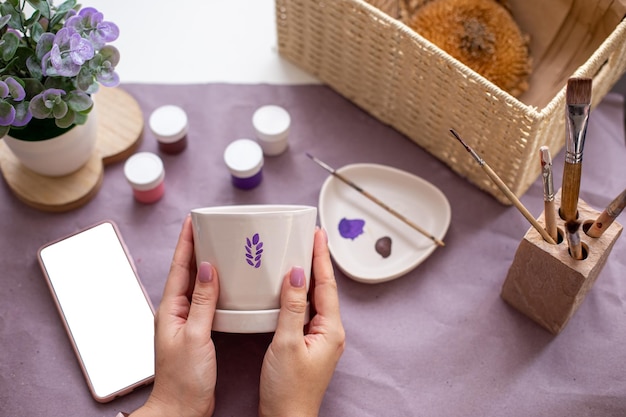 Womens hands hold a white flower ceramic pot on the table with craft tools