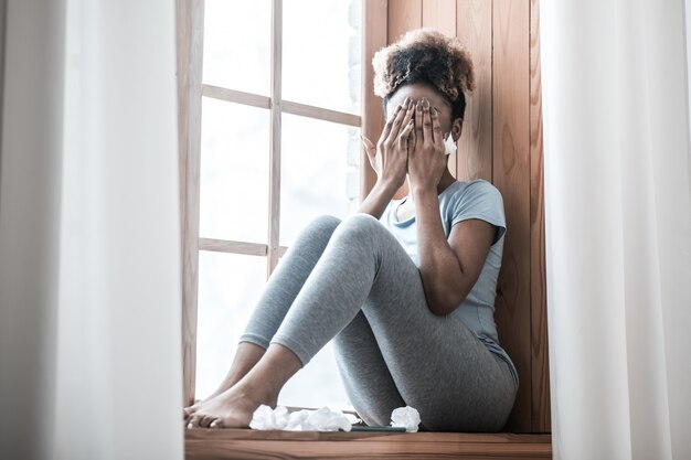 Womens tears. Young slender barefoot african american woman touching face with hands sitting on windowsill at home during day