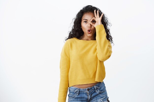 Wondered amused darkskinned girl with curly hairstyle wear yellow sweater folding lips gasping wow as checking out amazing cool promo look through circled fingers okay sign white background