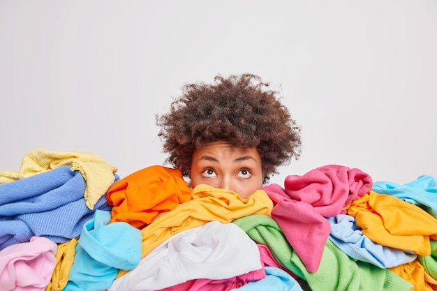 Wondered curly haired ethnic woman focused above surrounded by multicolored laundry cluttered with clothes collects clothing for recycling isolated over white wall. Organize your closet