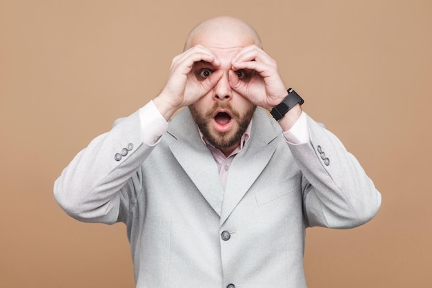 Wondered middle aged bald bearded businessman in classic light gray suit standing with binoculars gesture and looking at camera with shocked face. indoor studio shot, isolated on brown background.