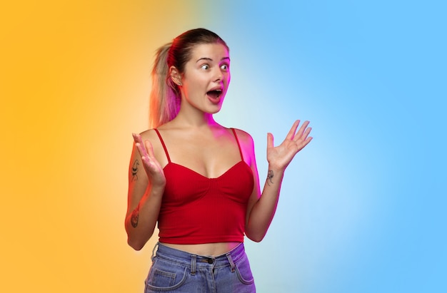 Wondered, shocked. Caucasian young woman's portrait on gradient studio background in neon. Beautiful female model in casual style. Concept of human emotions, facial expression, youth, sales, ad.