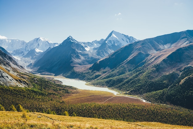 Wonderful alpine landscape with mountain lake and mountain river in valley with forest in autumn colors on background of snowy mountains silhouettes under blue sky. Beautiful mountain valley in autumn