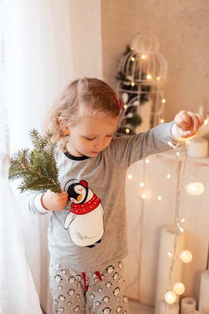 Wonderful beautiful little girl child in fashionable pajamas holding lights and Christmas tree branches