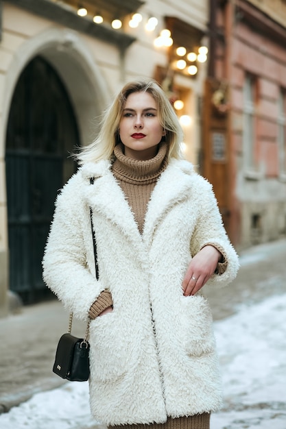 Meravigliosa modella bionda indossa cappotto invernale e borsa nera passeggiando per la città