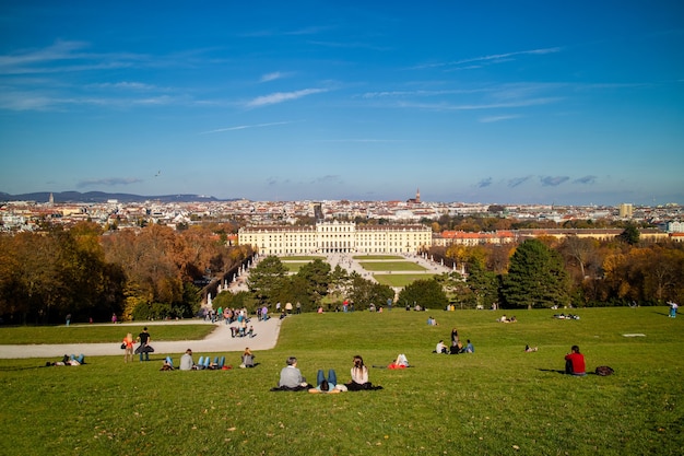 Wonderful landscape with view to schonbrunn palace in vienna, austria and green wide grass field with sitting and relaxing people on a ground on a blue sky background.