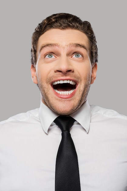 Wonderful news. Happy young man in shirt and tie smiling and looking up while standing against grey background