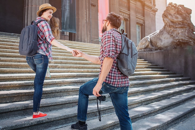 Wonderful picture of two tourists on steps outside on street. He stands lower and hold her hand. She looks back on man and smiles a bit. He has binoculars.