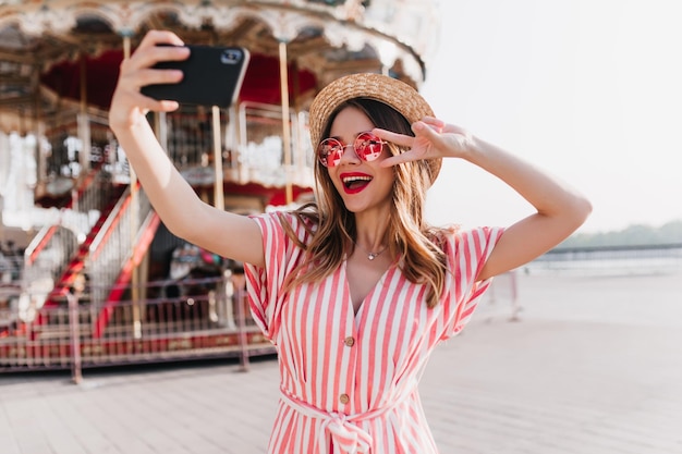 Meravigliosa signora magra con cappello che usa lo smartphone per selfie in un giorno d'estate foto all'aperto di una ragazza estatica in abito a righe che si fotografa nel parco divertimenti