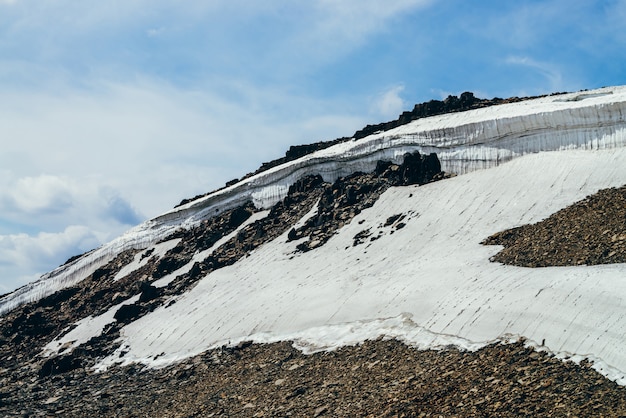 Wonderful view to small glacier on stony hill under blue sky with clouds. Beautiful alpine landscape with ice cornice on rock. Colorful mountain scenery on high altitude. Majestic highland nature.