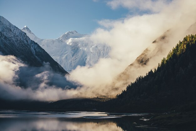 Wonderful view of snow-capped mountains above thick clouds in gold sunshine. Scenic landscape with alpine lake, silhouette of coniferous forest and high white-snow mountain wall among dense low clouds