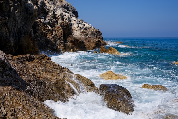 Wonderful views of the blue Mediterranean Sea. Sunny rocks, waves with foam and splashing water. The wave crashes into the rocks on the shore