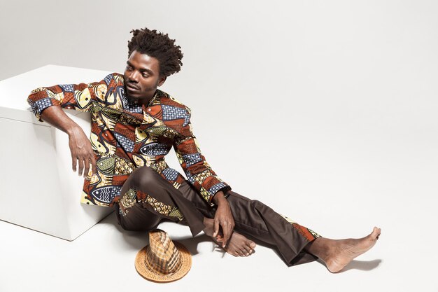 Wondering afro man in traditional national clothes sit on cube. Indoor, isolated on gray background