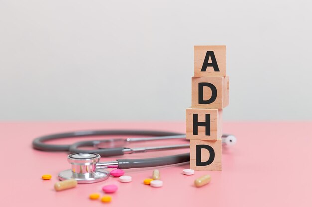 деревянный кубический блок Аббревиатура ADHD со стетоскопом и таблетками на розовом столе Дефицит внимания и гиперактивность ADHD концепция