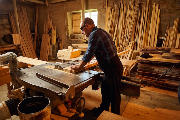 Wood cutting table with electric circular saw senior\
professional carpenter in uniform cutting wooden board at sawmill\
carpentry manufacturing sawing machine