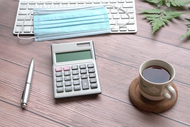 Wood table with keyboard face mask coffee pen calculator and\
green plants