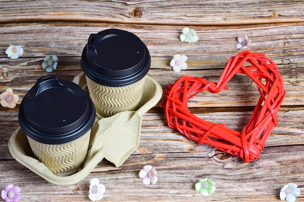 On a wooden background, two cups of coffee on the right and a large decorative wicker heart on the right. Small flowers are scattered around