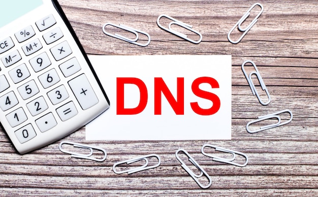 На деревянном фоне белый калькулятор, белые скрепки и белая карточка с текстом DNS Domain Name System. Вид сверху.