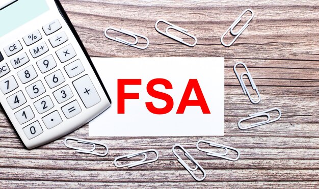 On a wooden background, a white calculator, white paper clips and a white card with the text FSA Flexible Spending Account. View from above.