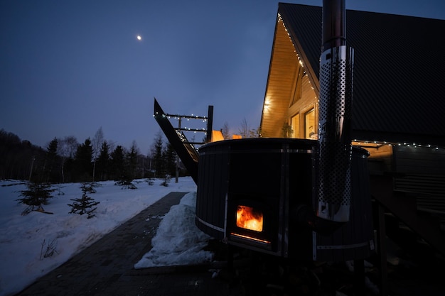 Wooden barrel hot tub in the terrace of the cottage at winter night Scandinavian bathtub with a fireplace to burn wood and heat water