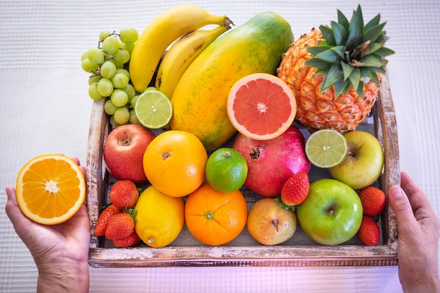 Wooden basket full of fresh fruit on a white background. Citrus fruits, papaya, pineapple, banana, pear, apple, strawberry, pomegranate, blueberry. Female hand holding half an orange