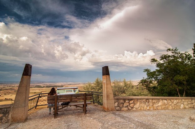 A wooden bench sits on a ledge overlooking a field and a mountain range.