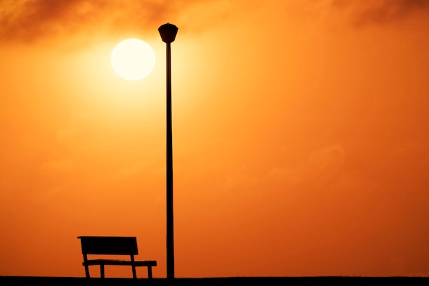 A Wooden bench and street lamp with a background view of a red sunset and a bright sun .